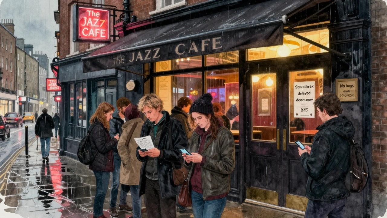 Patrons check their phones for updates outside The Jazz Cafe on a rainy London night, doors still closed.